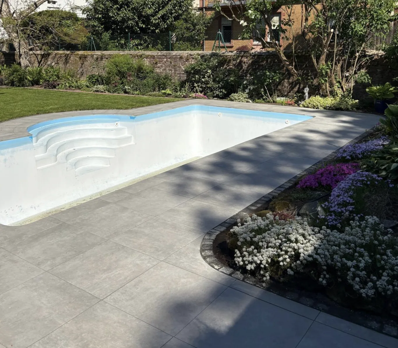 Pool mit grauer Plattenterrasse und geschwungener Beetkante mit blühenden Polsterstauden